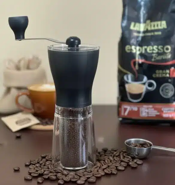 Manual coffee grinder filled with freshly ground coffee beans on a table, with whole coffee beans and espresso coffee in the background.